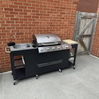 Assembled outdoor kitchen unit with grill, sink and storage against a brick wall